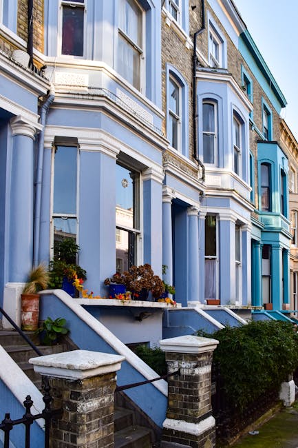 A row of Victorian terraced houses in Hammersmith with light blue and teal facades, featuring large bay windows and decorative white moldings. The houses are accessed by brick steps with white handrails leading up to the entrances, which are flanked by potted plants and flower boxes. The image shows a clear sky and front gardens with some greenery and paved areas. This setting illustrates the type of property where house removals and furniture transport should consider access issues, particularly during a home relocation or moving process. The historical architectural details and the urban environment highlight the challenges in loading and unloading items from properties with Victorian features, which is relevant to professional removals services provided by companies like Hammersmith Man and Van.