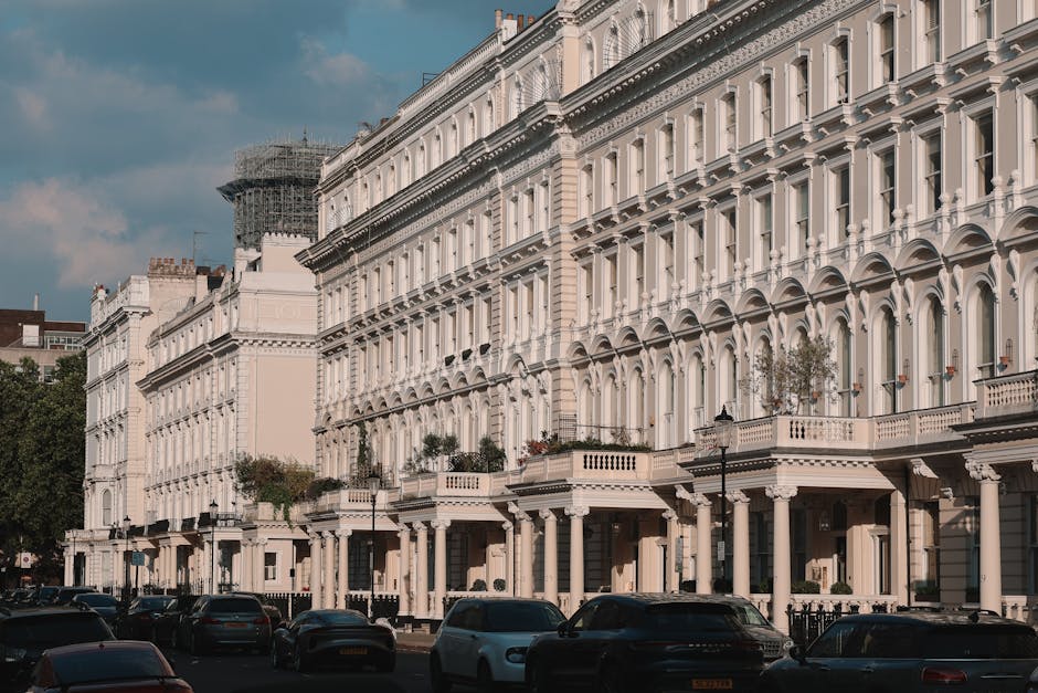 A row of white Victorian terrace houses with ornate architectural details, including arched windows, decorative cornices, and balcony railings, lined along a street with parked cars in the foreground. The buildings have large windows, some with flower pots on the balconies, and appear to be multiple stories tall. Scaffolding is visible on the left side of the image, indicating ongoing repairs or renovations. The scene is captured during daylight with a partly cloudy sky overhead. This setting illustrates typical urban residential architecture in Hammersmith, relevant to house removals and home relocation services provided by Hammersmith Man and Van. The street-level view shows the scale of furniture transport and packing logistics involved in moving within Victorian terraces, emphasizing the external environment where loading and unloading processes often take place during home moves.