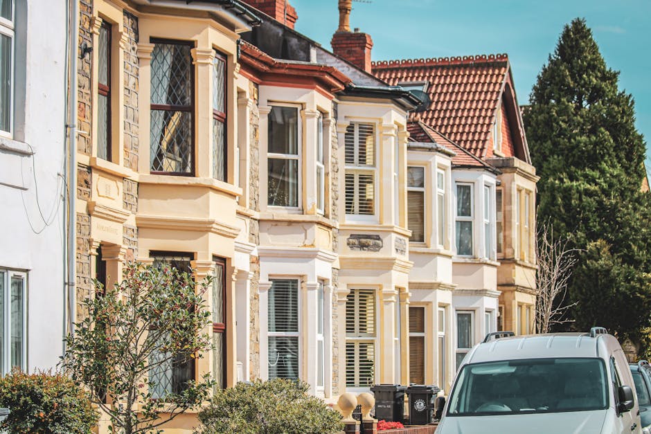 A row of Victorian terraced houses in Hammersmith with light blue and teal facades, featuring large bay windows and decorative white moldings. The houses are accessed by brick steps with white handrails leading up to the entrances, which are flanked by potted plants and flower boxes. The image shows a clear sky and front gardens with some greenery and paved areas. This setting illustrates the type of property where house removals and furniture transport should consider access issues, particularly during a home relocation or moving process. The historical architectural details and the urban environment highlight the challenges in loading and unloading items from properties with Victorian features, which is relevant to professional removals services provided by companies like Hammersmith Man and Van.