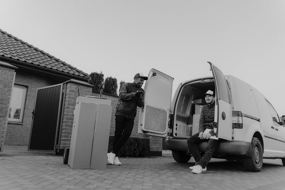 Two men involved in furniture transport during a home relocation, with one standing outside a residential property and the other sitting on the open rear door of a white cargo van. The man standing is dressed in a dark jacket, light trousers, and a cap, looking at his phone, while the seated man wears a jacket, jeans, and a hat, smiling toward the camera. Large cardboard boxes and packed items are placed on the driveway, ready for loading or unloading. Residential features such as a brick house with a tiled roof, a window, and a small garden are visible in the background. The scene shows a typical loading process, with furniture and boxes being moved into or out of the van, which is positioned near an entrance. The environment is well-lit, and the setting indicates professional removals, supporting home or furniture transport services offered by Hammersmith Man and Van, with attention to packing and moving logistics.