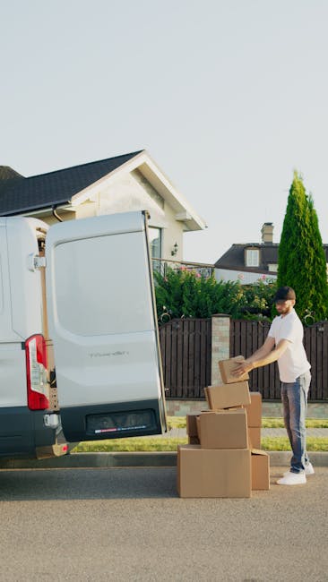A man standing next to an open white moving van parked on a residential street, with a row of cardboard boxes arranged on the pavement. The man is holding a box and appears to be preparing for home relocation activities such as packing or loading. The scene includes green hedges, tall trees, and houses with pitched roofs in the background. The van's interior is visible, showing space for furniture and packed items, while the weather is clear with bright daylight. This image illustrates the process of furniture transport and packing during a household move, relevant to professional removals services provided by Hammersmith Man and Van.