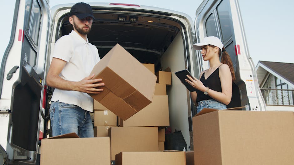 Two men involved in furniture transport during a home relocation, with one standing outside a residential property and the other sitting on the open rear door of a white cargo van. The man standing is dressed in a dark jacket, light trousers, and a cap, looking at his phone, while the seated man wears a jacket, jeans, and a hat, smiling toward the camera. Large cardboard boxes and packed items are placed on the driveway, ready for loading or unloading. Residential features such as a brick house with a tiled roof, a window, and a small garden are visible in the background. The scene shows a typical loading process, with furniture and boxes being moved into or out of the van, which is positioned near an entrance. The environment is well-lit, and the setting indicates professional removals, supporting home or furniture transport services offered by Hammersmith Man and Van, with attention to packing and moving logistics.