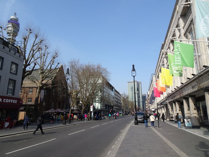 A wide street scene on Shepherds Bush Road during daytime, with a clear blue sky overhead. On the right side of the image, there is a large building with a series of colourful flags displaying logos such as 'H E A T S,' and other banners, indicating retail or commercial premises. Pedestrians are walking along the sidewalk, some carrying shopping bags or backpacks, and a few are waiting to cross at the intersection. On the left side, there are leafless trees lining the sidewalk, and several small outdoor market stalls with umbrellas, likely for local vendors, are visible near a building with a sign for a coffee shop. Moving vehicles, including cars and vans, are travelling along the multi-lane road, with some parked along the street. The surroundings feature a mix of modern and older architecture, with a tall building visible further down the road, suggesting the area is busy with foot traffic and vehicle movement typical of urban home relocation or furniture transport processes. This scene exemplifies the logistics and environment relevant to house removals and moving services, with a focus on outdoor staging and busy street activity.