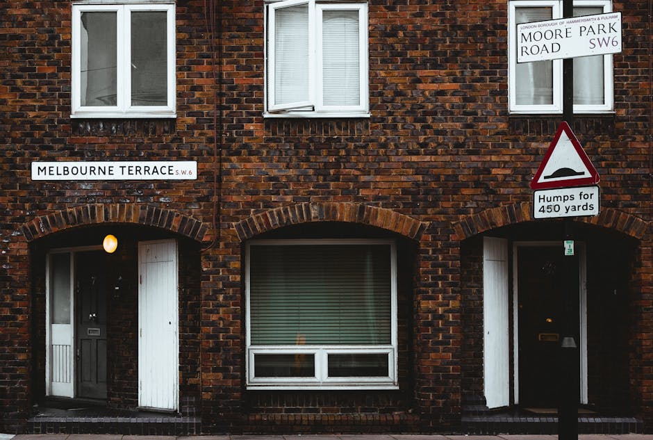 Photograph of a brick residential building at the corner of Melbourne Terrace and Moore Park Road in Hammersmith, displaying two upper windows with white frames and two ground-floor entrances with white doors, one of which has a window with blinds. The building's exterior features a traditional red brick façade with arched brickwork above the doorways. To the right of the building, there are street signs indicating the location as Melbourne Terrace SW6 and Moore Park Road SW6, along with a triangular warning sign for bumps or uneven road surfaces, stating 'Humps for 450 yards.' The image is taken during daylight, with natural light illuminating the scene. This setting relates to house removals and furniture transport, with the street signs and residential environment supporting the context of moving services in Fulham, Hammersmith, offered by Hammersmith Man and Van.