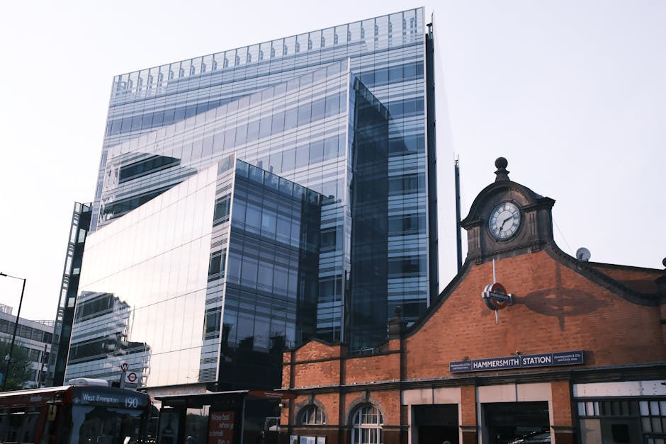 An urban scene featuring a modern, glass-fronted office building with reflective windows towering above a historic brick building with a clock face and a station sign reading 'Hammersmith Station.' In