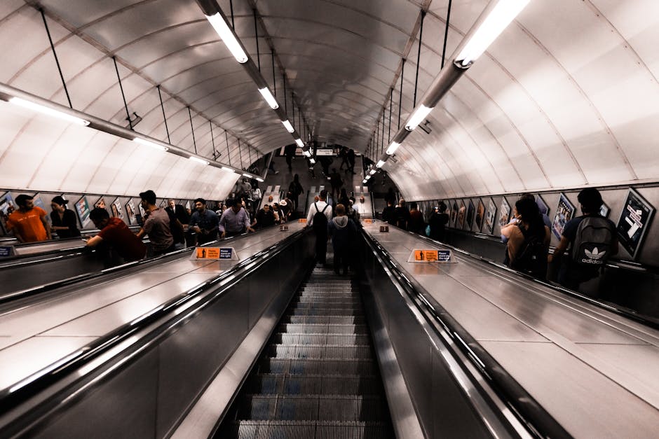 Inside a busy underground station with an escalator descending towards a platform where numerous people are waiting and walking. The station features a curved, white ceiling with linear fluorescent lighting fixtures. On either side of the escalator, there are metallic handrails and safety barriers, with framed posters or advertisements attached to the wall panels. The crowd includes individuals carrying backpacks, handbags, and luggage, reflecting a typical transit environment. The ambient lighting is bright and even, emphasizing the sleek, modern design of the station. This setting highlights the context of urban mobility, potentially related to home relocation or transport logistics, where moving companies like Hammersmith Man and Van may assist with furniture transport or packing and moving services, especially in busy public transit locations.