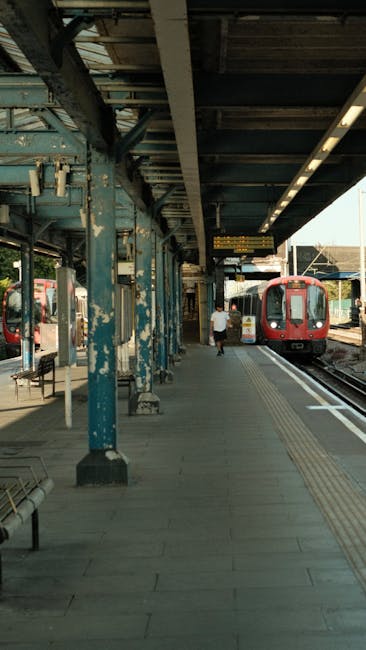 Inside a railway station platform with a covered roof supported by weathered blue-painted metal columns, some with peeling paint, and a grey tiled floor featuring tactile paving strips for visually impaired passengers. There is a red passenger train stopped at the platform, with windows and doors visible. A single person in casual clothing is walking towards the train, carrying a small bag. To the left, empty benches are positioned along the platform. The station appears to be well-lit with natural light coming from the open side where the trains are parked. Visible on the platform are digital information boards and safety markings, with some equipment such as a trolley nearby. This scene illustrates a typical setting for home relocation tasks involving packing, transporting, and loading furniture or boxes onto trains, reflecting the importance of efficient moving logistics... Hammersmith Man and Van.