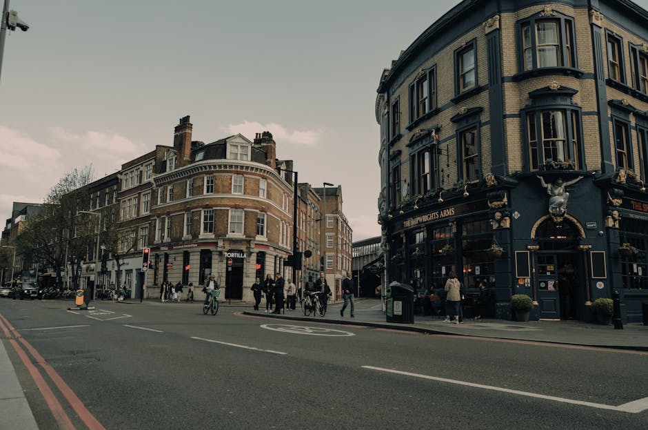 A city street scene in Hammersmith featuring a mix of historic and modern buildings, including a rounded corner pub with ornate decorations and a navy blue facade named 'The Star Wights Arms' on the ground floor. Pedestrians are walking along the sidewalk, some crossing the intersection, while cyclists navigate through the area. Several cars and a van are parked along the curb, with a traffic light visible at the corner. The road displays red double lines on the edge, indicating parking restrictions, and the surroundings are illuminated by natural daylight, capturing an active urban environment suitable for house removals and furniture transport services offered by Hammersmith Man and Van. This scene illustrates the typical setting for loading or unloading during a home relocation in central Hammersmith.