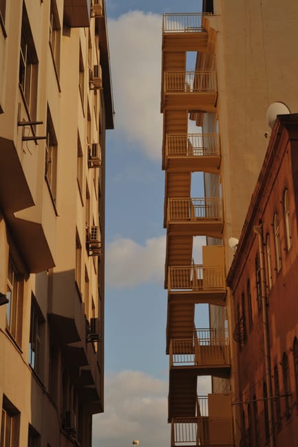 Photograph taken from ground level showing the external fire escape staircase of a tall, beige-colored building on the right side, consisting of several narrow, open metal balconies with railings, extending vertically along the facade. To the left, part of another building with multiple windows and air conditioning units attached beneath some of the window frames is visible. The sky is partly cloudy with patches of blue, and the scene is illuminated by natural daylight. This image captures the typical urban residential environment relevant to house removals and furniture transport, illustrating space constraints often faced during home relocations, especially when navigating narrow staircases and external access points, as discussed in W6 man and van tips for narrow staircases and bays at Hammersmith.