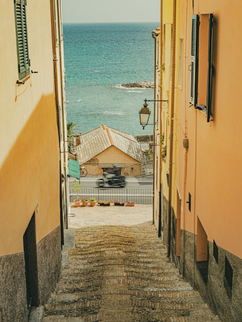 A narrow stone stairway leading down from a residential area with yellow and orange painted exterior walls on either side, featuring closed window shutters and a wall-mounted lantern. The staircase is outdoors, with visible textured steps, and opens to a street scene with a row of potted plants along the pavement. Beyond the street, a small building with a tiled roof is visible, situated near the coastline, with the sea extending to the horizon in the background. The scene is illuminated by natural daylight, highlighting the vibrant wall colours and the scenic coastal view. This setting exemplifies the typical environment encountered during house removals or relocation tasks near waterfront areas, where careful handling of furniture and packing materials might be required for home relocation or furniture transport by professionals such as Hammersmith Man and Van.