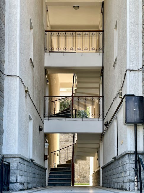 Photograph taken from ground level showing the external fire escape staircase of a tall, beige-colored building on the right side, consisting of several narrow, open metal balconies with railings, extending vertically along the facade. To the left, part of another building with multiple windows and air conditioning units attached beneath some of the window frames is visible. The sky is partly cloudy with patches of blue, and the scene is illuminated by natural daylight. This image captures the typical urban residential environment relevant to house removals and furniture transport, illustrating space constraints often faced during home relocations, especially when navigating narrow staircases and external access points, as discussed in W6 man and van tips for narrow staircases and bays at Hammersmith.